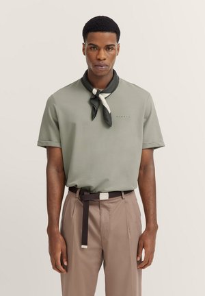 Olivgrünes Poloshirt mit kurzen Ärmeln, kombiniert mit einem schwarz-weißen Halstuch, beigem Hosen und einem braunen Gürtel mit einer Metallschnalle.