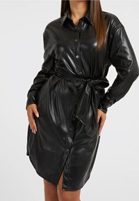 Femme portant une robe en faux cuir noir à boutons, à manches longues et avec une ceinture nouée à la taille, se tenant devant un fond clair.