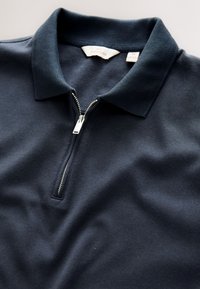 Polo bleu marine à fermeture éclair avec col côtelé, partiellement zippé, montrant une étiquette de taille moyenne à l'intérieur de l'encolure.