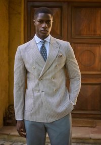 Giacca da completo doppiopetto beige chiaro con righe verticali, abbinata a una camicia dress light blue e una cravatta con motivo. Visibile un notevole fazzoletto da taschino.