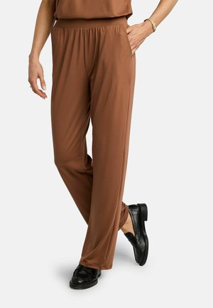 Mujer vistiendo pantalones marrones de corte holgado con cintura elástica y zapatos negros sin cordones, con una mano en el bolsillo, sobre fondo blanco.