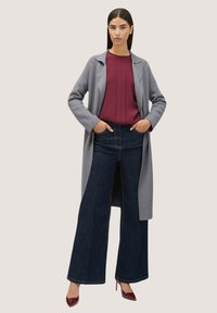 Cappotto lungo grigio sopra una blusa bordeaux, abbinato a jeans a gamba larga blu scuro e tacchi bordeaux. Tessuto morbido, design su misura e vestibilità strutturata.