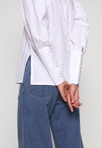 Camicia bianca a maniche lunghe con maniche a sbuffo, con dettagli in perle sui polsini, fessure laterali e una texture morbida. Indossata con jeans blu.