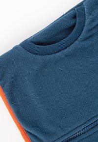 Blaues Fleece-Sweatshirt mit geripptem Rundhalsausschnitt und orangefarbenem seitlichem Einsatz. Verfügt über einen Reißverschlussdetails auf der unteren rechten Seite. Weiche Textur.
