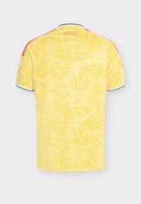 Gul fotbollströja från Colombia med subtila mönster av fjärilsvingar, röda ränder på axlarna och blå kantning på ärmar och krage.