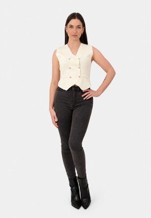 Frau mit langen dunklen Haaren trägt ärmellose cremefarbene Weste, schwarze High-Waist Skinny Jeans und schwarze spitze Stiefel, steht mit der Hand in der Hüfte.
