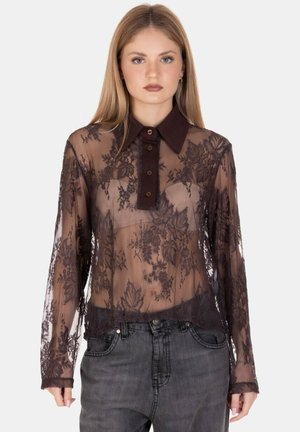 Giovane donna che indossa una blusa nera in pizzo trasparente con motivi floreali sopra un reggiseno beige, abbinata a jeans neri sbiaditi su uno sfondo semplice.