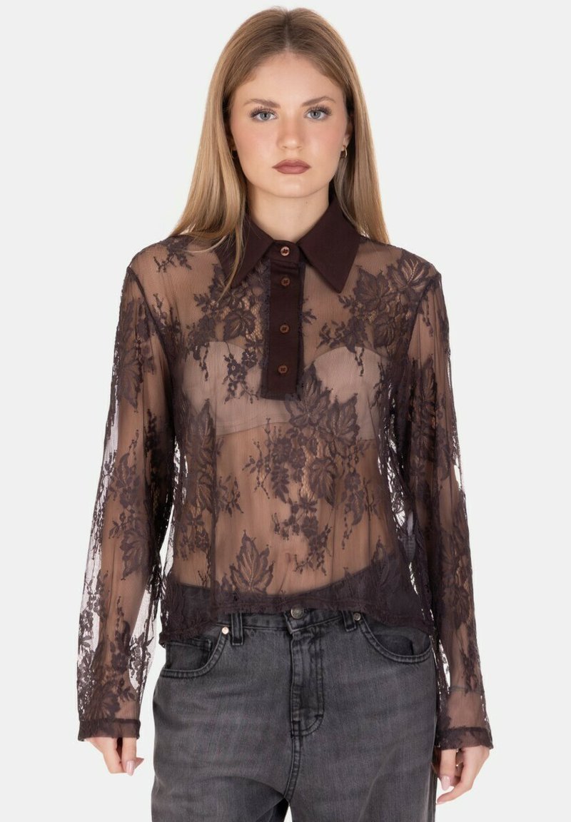 Giovane donna che indossa una blusa nera in pizzo trasparente con motivi floreali sopra un reggiseno beige, abbinata a jeans neri sbiaditi su uno sfondo semplice.