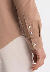 Chemise marron à boutons, présentant une texture lisse, des boutons blancs et un ourlet légèrement incurvé. Les poignets ont un design pointu et trois fermetures à boutons.