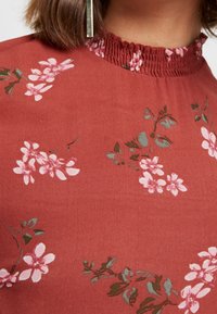 Tæt på en person iført en rustfarvet blomstret top med pink blomster og grønne blade, der har en rynket høj hals og en vertikal metallisk øresmykke.