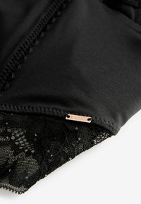 Schwarzes Stoffoberteil mit filigranem Spitzenbesatz und Pailletten am Saum sowie einem kleinen metallischen Logo-Tag. Glatte Textur und taillierte Passform.