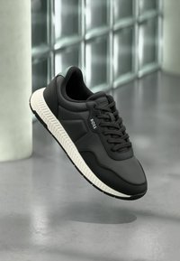 Μαύρο sneaker BOSS με λευκή σόλα, λεπτομέρειες από δίχτυ και δέρμα, δεμένο με κορδόνια, εμφανίζεται στον αέρα μπροστά σε μοντέρνο φόντο από γυάλινα τούβλα.