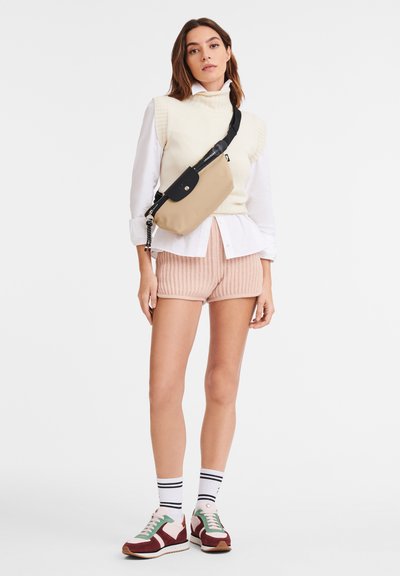 Beige crossbody bag with black strap, cream ribbed sweater over white shirt, light pink shorts, and multicolored sneakers with striped socks.