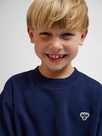 Jonge jongen met blond haar die lacht, draagt een marineblauw truitje met een klein wit en blauw logo op de borst, tegen een effen lichte achtergrond.