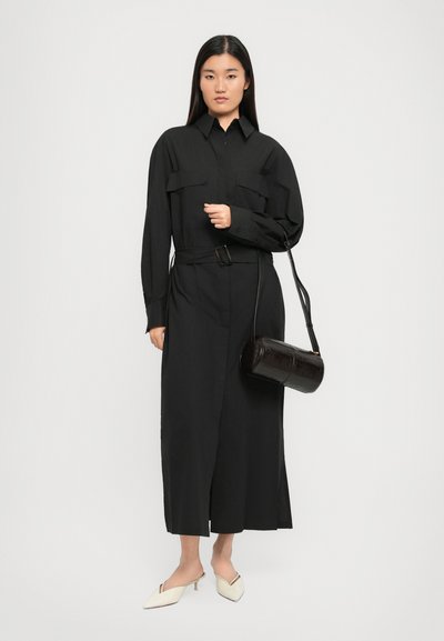 Black long dress with button-up collar, chest pockets, and a belt. Paired with a shiny black cylindrical bag and cream pointed shoes.