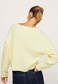 Maglione lavorato a maglia di un giallo chiaro con texture a coste, ampia scollatura a barca, spalle sagomate e vestibilità leggermente accorciata. Indossato con jeans scuri.