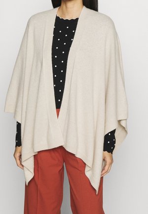 Frau trägt einen beigen Poncho mit offenem Vorderteil über einem schwarzen, gepunkteten Oberteil und rostfarbenen Hosen, steht vor einem schlichten Hintergrund.