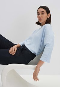 Hellblau gestreifter Pullover mit Dreiviertelärmeln, kombiniert mit dunklen Hosen. Das Modell lehnt lässig auf einem weißen skulpturalen Stuhl.