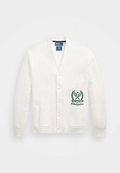 Cardigan blanc avec un col en V, cinq boutons, deux poches avant et un logo de Wimbledon vert brodé sur le côté gauche. Texture de tissu douce.