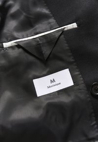 Juodo audinio striukės vidus, kuriame yra balta etiketė su "Matinique" tekstu, apsupta kontrastingo rašto, mygtuko ir kišenės detalėmis.