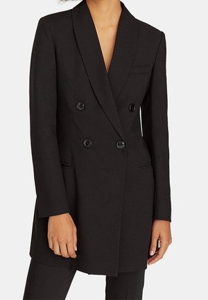 Vrouw draagt een lange zwarte dubbelgeknoopte blazer met vier knopen en twee voorzakken, gecombineerd met een zwarte broek.