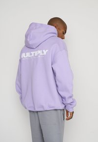 Sudadera de color púrpura claro con una capucha amplia, puños y dobladillo acanalados, con un logotipo gráfico blanco impreso en la parte posterior. Combinada con pantalones grises.