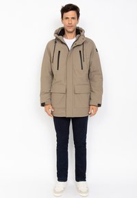 Chaqueta aislante beige con capucha, cremallera frontal y dos bolsillos en el pecho. Presenta cremalleras negras y cordones ajustables. Llevada con jeans oscuros.