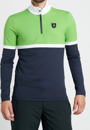 Mężczyzna noszący longsleeve z blokami kolorów zielonego, białego i granatowego oraz przednim zamkiem, stoi z rękami luźno opuszczonymi.