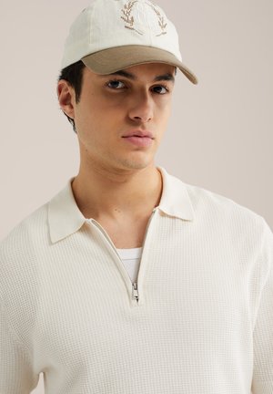 Mladić u bež i bijeloj baseball kapici te teksturiranoj majici svijetle boje sa zatvaračem i ovratnikom, na neutralnoj pozadini.