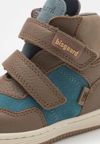 Chaussure enfant en cuir marron et en daim bleu avec des sangles Velcro, surface texturée et accents beiges. Présente une étiquette de marque sur le côté.