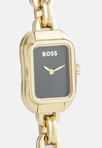 Montre rectangulaire dorée avec un cadran gris foncé, au design minimaliste, bracelet maillé et logo "BOSS" en centre.
