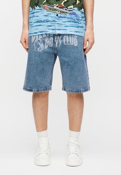 Férfi, aki középhosszú kék farmer rövidnadrágot visel, elöl "Billionaire Boys Club" felirattal és űrhajós grafikával, fehér sportcipővel és zoknival párosítva.