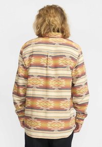 Persoon met schouderlang blond haar, gekleed in een langemouwenshirt met horizontale strepen en geometrische patronen in warme kleuren, gezien van achteren.