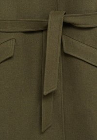 Detailní záběr na olivově zelený látkový kabát s páskem svázaným do uzlu a klopovými kapsami po obou stranách.