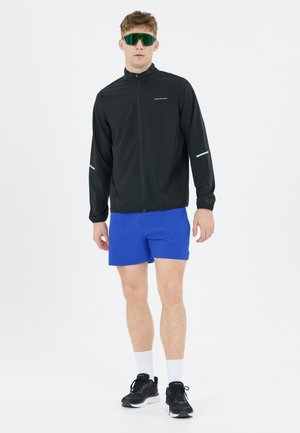 Man staat naar voren gericht, draagt reflecterende zonnebril, een zwart jack met rits, felblauwe korte broek, witte sokken en zwarte sneakers.
