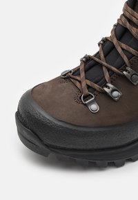Brun utendørs støvel med teksturert semsket lær, gummiforlenger ved tåen, metalløyler og mørke snører. Har en robust såle med godt grep.