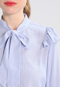 Hellblaue, gestreifte Bluse mit einem Schleifenkragen und rüschend Akzenten an den Schultern. Glatte Stofftextur mit vertikalen weißen Streifen.