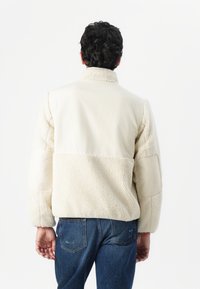 Kremfarget, teksturert fleecejakke med høy krage, kontrasterende glatte stoffseksjoner og lange ermer, tilsatt blå jeans.