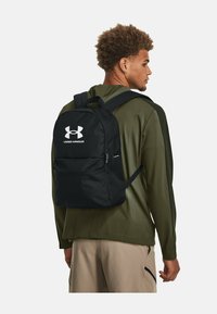 Čierny plátkový batoh s veľkým logom Under Armour, polstrovanými popruhmi a predným vreckom na zips, nosí ho osoba v olivovom saku.