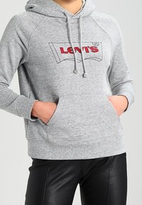 Sudadera gris con un bolsillo tipo canguro, logo rojo de "LEVI'S" en la parte frontal, puños acanalados y capucha con cordones. Combinada con pantalones de cuero negros.