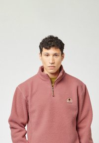 Fleece pullover in zacht roze met een kwart rits, met een gestructureerd oppervlak en een circulair logo op de borst. De binnenlaag is gestreept groen.