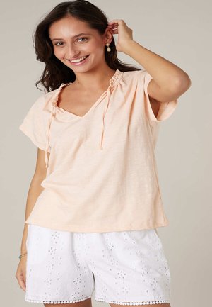 Femme souriante ajustant sa boucle d'oreille, portant un haut pêche à manches courtes et un short blanc en dentelle œillet, sur un fond uni.