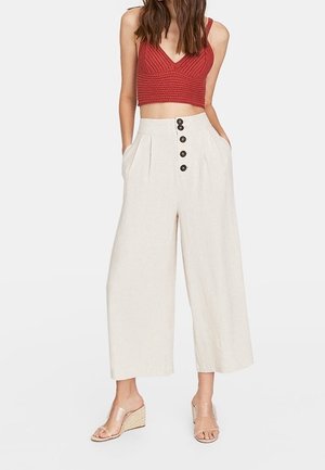 Vrouw draagt een rood gebreide crop top, hooggetailleerde beige broek met wijde pijpen en zwarte knopen, en transparante sleehak sandalen.