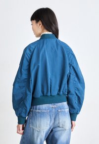 Chaqueta bomber color verde azulado con un corte holgado, puños elásticos y cinturilla. Presenta una textura suave y mangas fruncidas, combinadas con jeans de un azul claro.