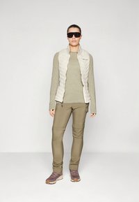 Kerge kreemjas puffy vest sobitatud õhukese oliivrohelise pika varrukaga särgi, oliivroheliste kitsaste pükstega ja tumeda halli matkajalatsitega.