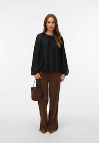 Blusa nera con scollatura arricciata, maniche lunghe. Pantaloni larghi marroni. La modella tiene una piccola borsa marrone, indossando scarpe beige a punta.