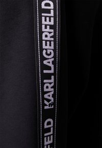 Zwarte kledingstuk met een textuurstof en een opvallende verticale band met een wit logo die "KARL LAGERFELD" leest. Eenvoudig ontwerp en kleurencontrast.