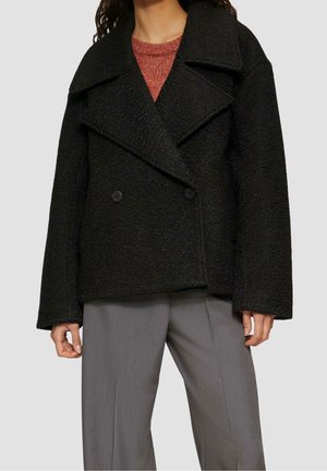 Person wearing a black textured double-breasted coat over a rust-colored sweater and gray wide-leg pants against a plain background.