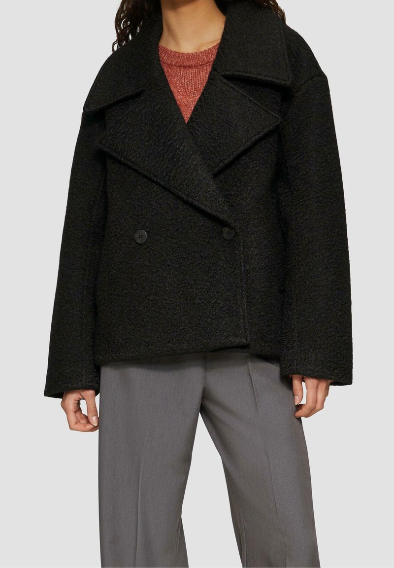 Personne portant un manteau noir texturé à double boutonnage par-dessus un pull couleur rouille et un pantalon gris à jambes larges sur un fond uni.