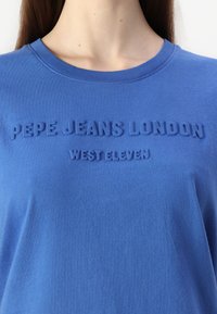 Mėlynas medvilninis marškinėlis su apvaliu iškirpimu. Priekyje yra iškilęs užrašas "PEPE JEANS LONDON WEST ELEVEN". Lygus paviršius.